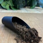 Zbliżenie na szarą lub zieloną plastikową łopatkę do nabierania podłoża, leżącą obok opakowania granulatu Lechuza PON. Łopatka ma wąski, wyprofilowany kształt i wygodny uchwyt.